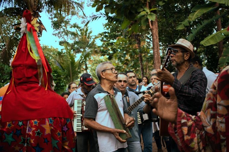 Foliões de reis cantam e tocam sanfona e viola ao lado de um palhaço de máscara vermelha.