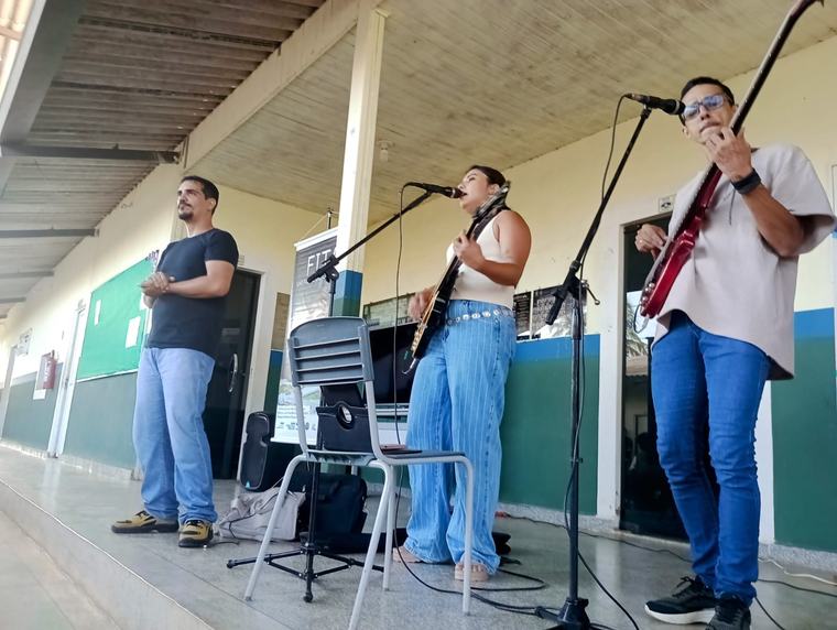 Três integrantes do coletivo tocam e cantam no pátio coberto de uma escola.