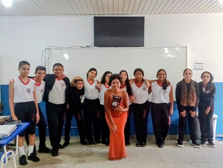 Turma de alunos posa numa sala de aula ao lado de uma artista de vestido laranja, que segura um livro.