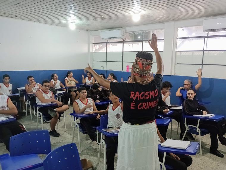Artista com máscara e camiseta com a frase “Racismo é crime” se apresenta numa sala de aula enquanto alunos levantam a mão.