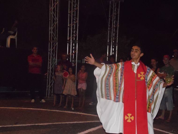 Ator de túnica colorida e estola vermelha se apresenta à noite, ao ar livre, diante do público.
